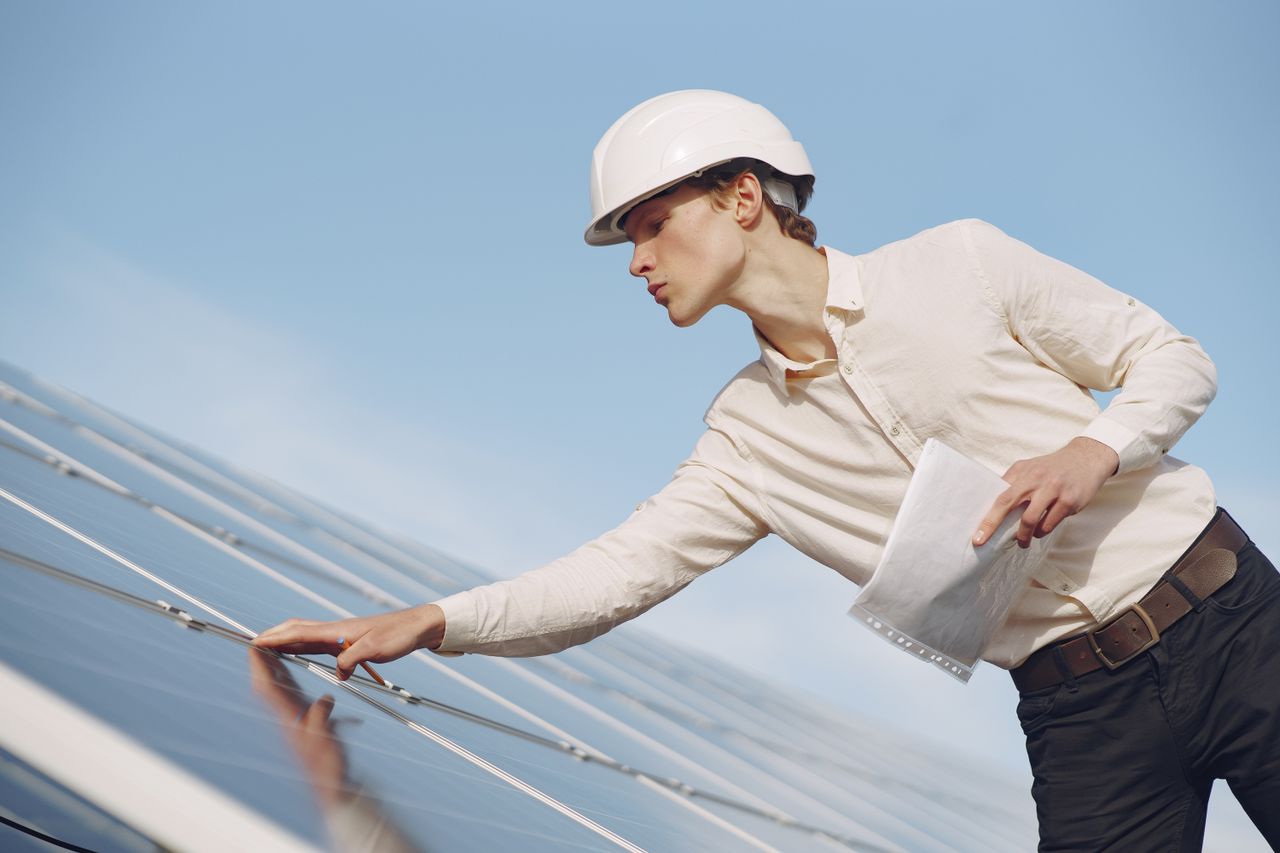 gallery-4 Low angle of young male inspector with documents in formal wear and hardhat standing against solar panels and checking details of construction