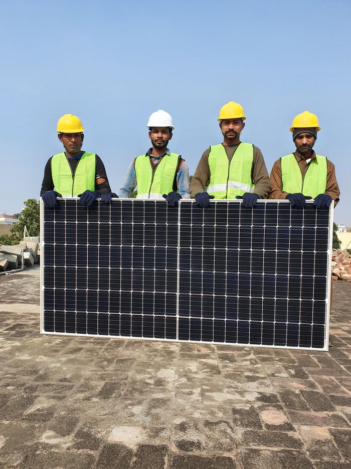 gallery-1 Group of construction workers holding a solar panel on a sunny day, wearing safety gear.